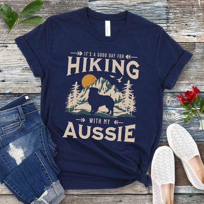 A navy t-shirt with a vintage-style graphic print of mountains, an Australian Shepherd silhouette, and the text 'Hiking with my Aussie'. The shirt is presented on a wooden surface with a pair of blue jeans and white shoes in the background.