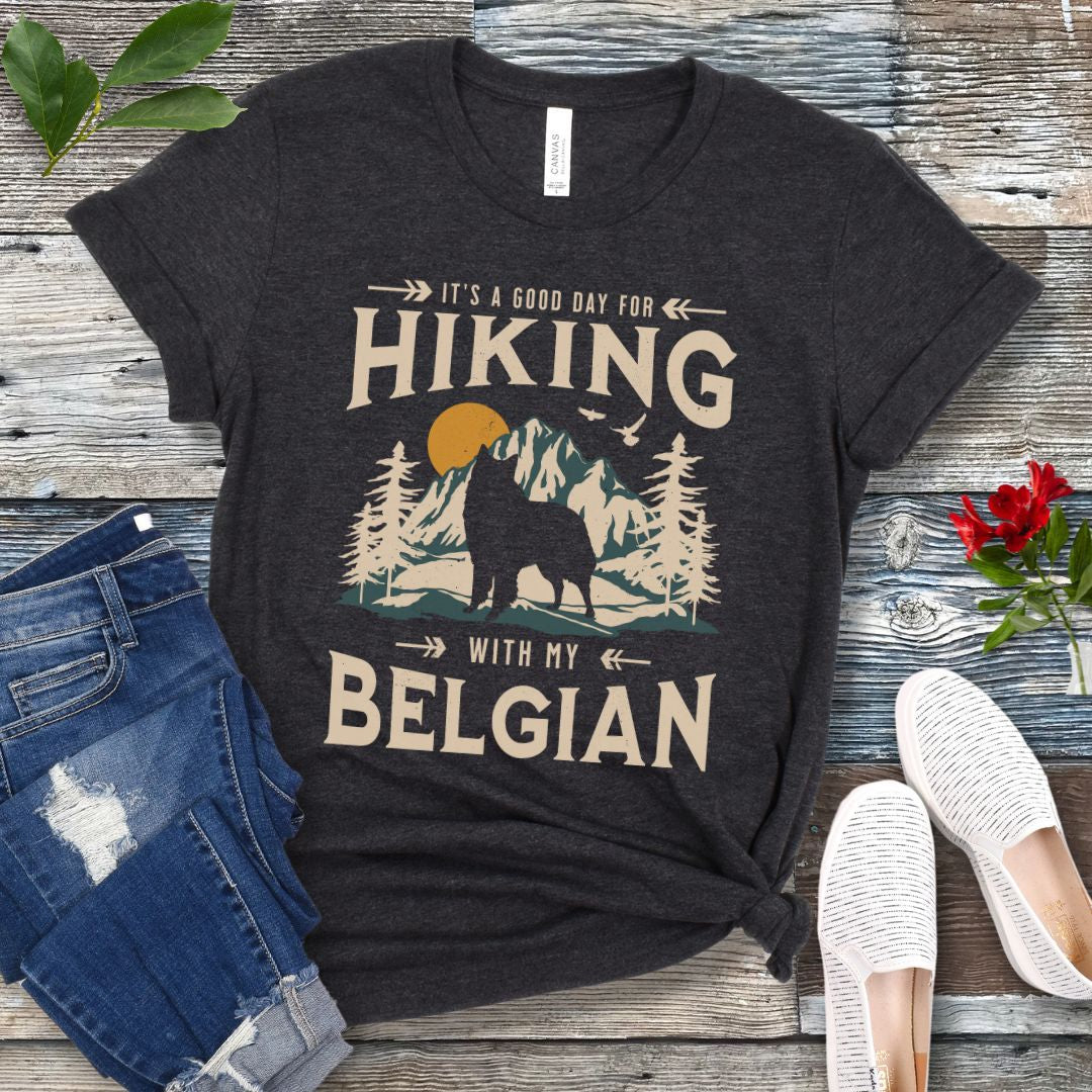 A dark grey t-shirt with a vintage-style graphic print of mountains and a Belgian Sheepdog and the text 'Hiking with my Belgian'. The shirt is presented on a wooden surface with a pair of blue jeans and white shoes in the background.