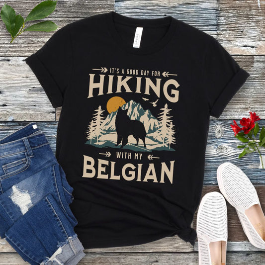 A black t-shirt with a vintage-style graphic print of mountains and a Belgian Sheepdog and the text 'Hiking with my Belgian'. The shirt is presented on a wooden surface with a pair of blue jeans and white shoes in the background.
