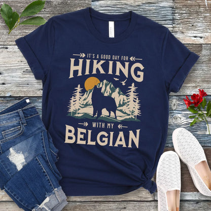 A navy blue t-shirt with a vintage-style graphic print of mountains and a Belgian Sheepdog and the text 'Hiking with my Belgian'. The shirt is presented on a wooden surface with a pair of blue jeans and white shoes in the background.