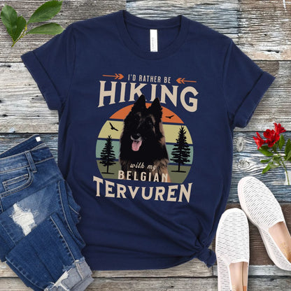 A navy Bella Canvas 3001 unisex short sleeve T-shirt with a graphic print of a Belgian Tervuren dog and the text 'I'd Rather Be Hiking With My Belgian Tervuren'. The shirt is presented on a wooden surface with a pair of blue jeans and white shoes in the background.