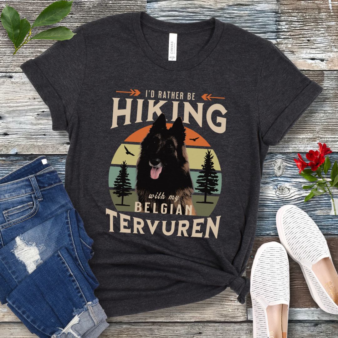 A dark grey Bella Canvas 3001 unisex short sleeve T-shirt with a graphic print of a Belgian Tervuren dog and the text 'I'd Rather Be Hiking With My Belgian Tervuren'. The shirt is presented on a wooden surface with a pair of blue jeans and white shoes in the background.