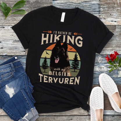 A black Bella Canvas 3001 unisex short sleeve T-shirt with a graphic print of a Belgian Tervuren dog and the text 'I'd Rather Be Hiking With My Belgian Tervuren'. The shirt is presented on a wooden surface with a pair of blue jeans and white shoes in the background.