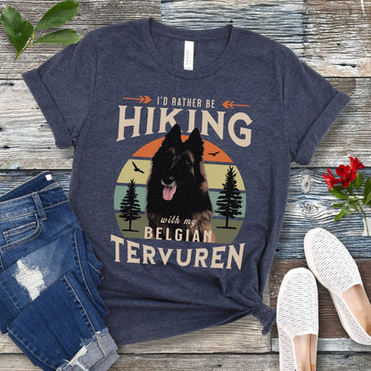 A heather navy Bella Canvas 3001 unisex short sleeve T-shirt with a graphic print of a Belgian Tervuren dog and the text 'I'd Rather Be Hiking With My Belgian Tervuren'. The shirt is presented on a wooden surface with a pair of blue jeans and white shoes in the background.