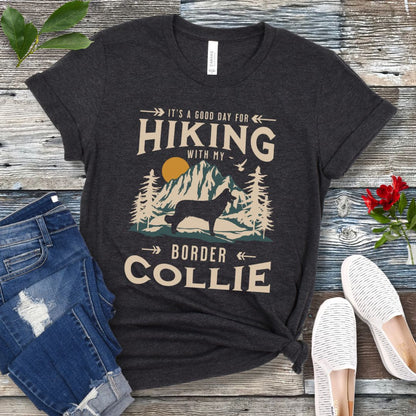 A dark grey t-shirt with a vintage-style graphic print of mountains and a Border Collie and the text 'Hiking with my Border Collie'. The shirt is presented on a wooden surface with a pair of blue jeans and white shoes in the background.