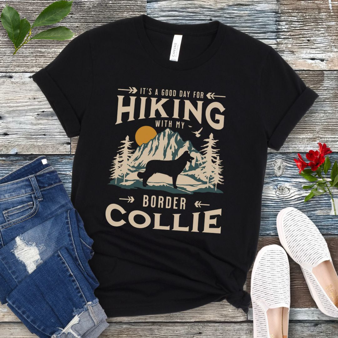 A black t-shirt with a vintage-style graphic print of mountains and a Border Collie and the text 'Hiking with my Border Collie'. The shirt is presented on a wooden surface with a pair of blue jeans and white shoes in the background.