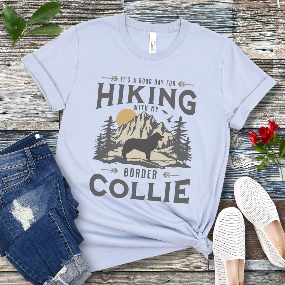 Light blue t-shirt for Border Collie lovers with vintage style mountain graphic and Border Collie silhouette with the text 'It's a Good Day For Hiking With My Border Collie'. Shown on a wooden surface with jeans and shoes.
