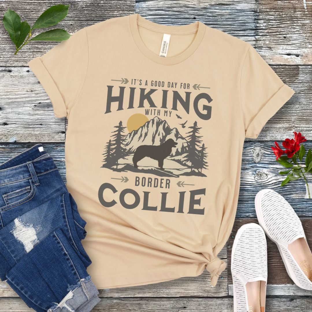 Cream colored t-shirt for Border Collie lovers with vintage style mountain graphic and Border Collie silhouette with the text 'It's a Good Day For Hiking With My Border Collie'. Shown on a wooden surface with jeans and shoes.
