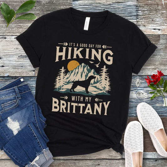 A black t-shirt with a vintage-style graphic print of mountains, a Brittany Spaniel silhouette, and the text 'Hiking with my Brittany'. The shirt is presented on a wooden surface with a pair of blue jeans and white shoes in the background.