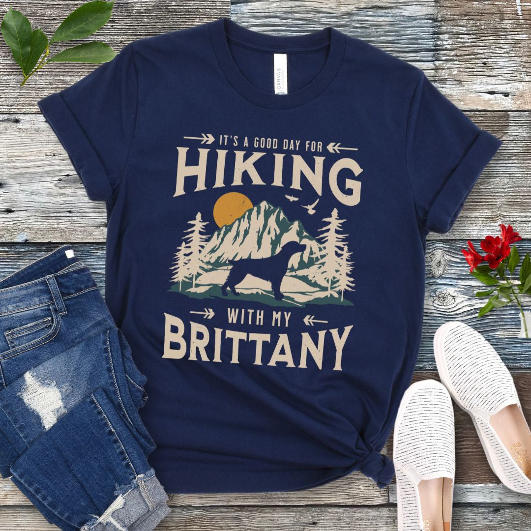 A navy blue t-shirt with a vintage-style graphic print of mountains, a Brittany Spaniel silhouette, and the text 'Hiking with my Brittany'. The shirt is presented on a wooden surface with a pair of blue jeans and white shoes in the background.