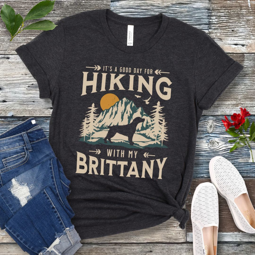 A dark grey t-shirt with a vintage-style graphic print of mountains, a Brittany Spaniel silhouette, and the text 'Hiking with my Brittany'. The shirt is presented on a wooden surface with a pair of blue jeans and white shoes in the background.