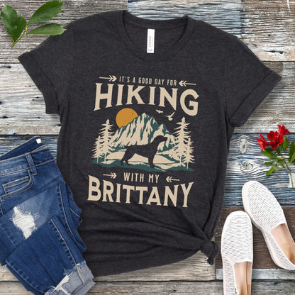 A dark grey t-shirt with a vintage-style graphic print of mountains, a Brittany Spaniel silhouette, and the text 'Hiking with my Brittany'. The shirt is presented on a wooden surface with a pair of blue jeans and white shoes in the background.