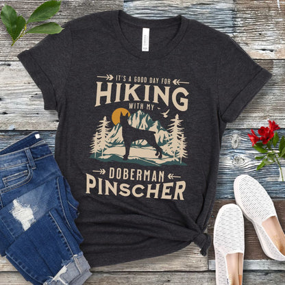A dark grey t-shirt with a vintage-style graphic print of mountains and a Doberman and the text 'Hiking with my Doberman Pinscher'. The shirt is presented on a wooden surface with a pair of blue jeans and white shoes in the background.
