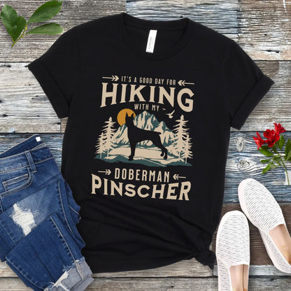 A black t-shirt with a vintage-style graphic print of mountains and a Doberman and the text 'Hiking with my Doberman Pinscher'. The shirt is presented on a wooden surface with a pair of blue jeans and white shoes in the background.