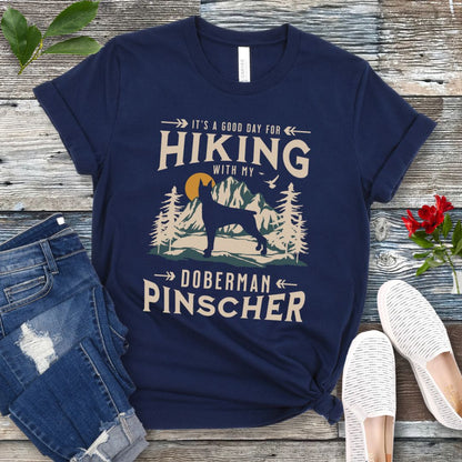 A navy t-shirt with a vintage-style graphic print of mountains and a Doberman and the text 'Hiking with my Doberman Pinscher'. The shirt is presented on a wooden surface with a pair of blue jeans and white shoes in the background.