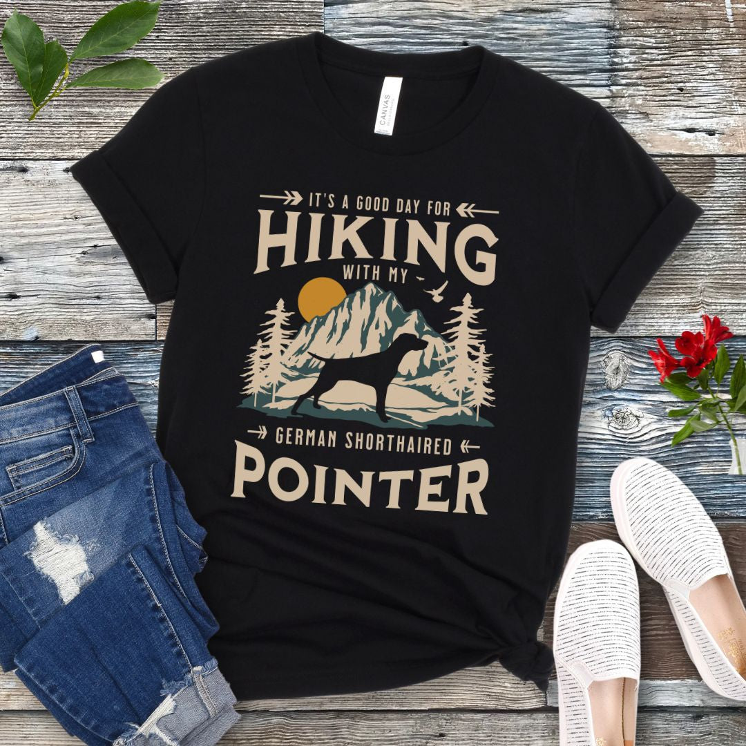 A black t-shirt with a vintage-style graphic print of mountains, a German Shorthaired Pointer silhouette, and the text 'Hiking with my German Shorthaired Pointer'. The shirt is presented on a wooden surface with a pair of blue jeans and white shoes in the background.