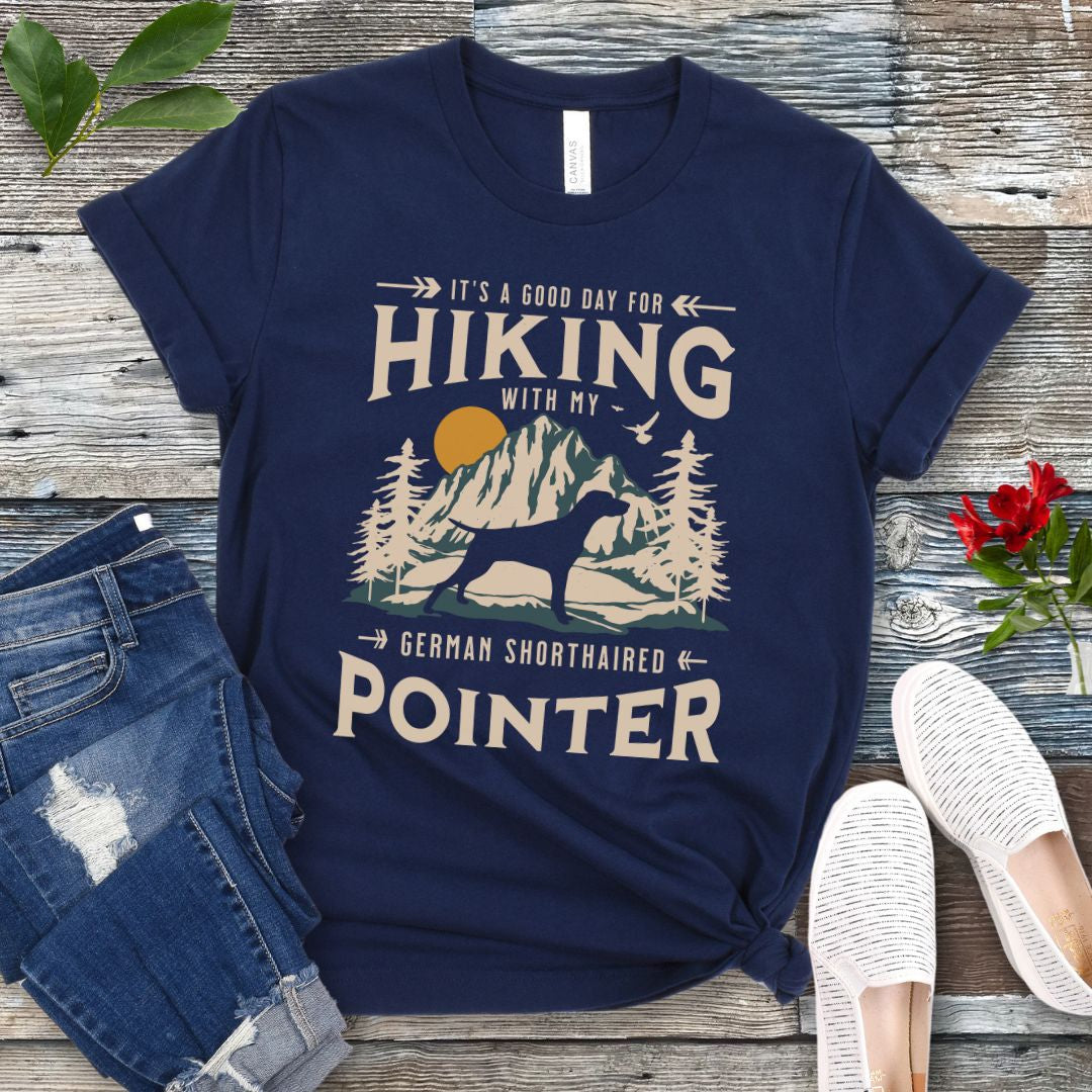 A navy blue t-shirt with a vintage-style graphic print of mountains, a German Shorthaired Pointer silhouette, and the text 'Hiking with my German Shorthaired Pointer'. The shirt is presented on a wooden surface with a pair of blue jeans and white shoes in the background.