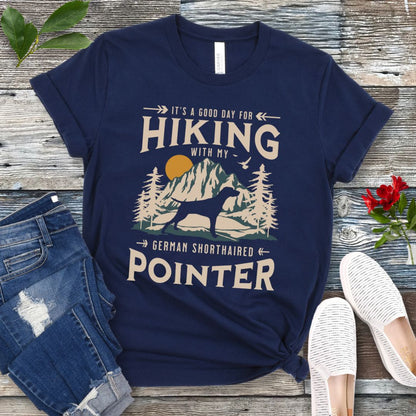 A navy blue t-shirt with a vintage-style graphic print of mountains, a German Shorthaired Pointer silhouette, and the text 'Hiking with my German Shorthaired Pointer'. The shirt is presented on a wooden surface with a pair of blue jeans and white shoes in the background.