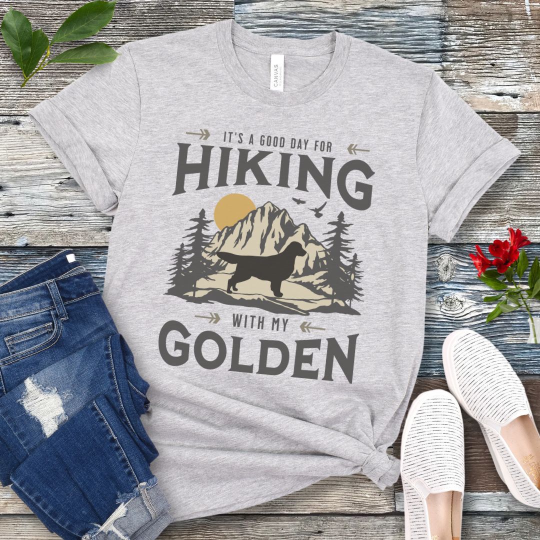Grey t-shirt with vintage mountain scene, Golden Retriever silhouette and 'Hiking with my Golden' text, presented on a wooden surface with jeans and shoes.
