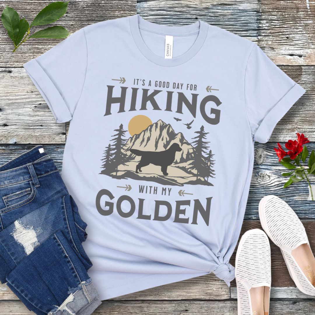 Light blue t-shirt with vintage mountain scene, Golden Retriever silhouette and 'Hiking with my Golden' text, presented on a wooden surface with jeans and shoes.