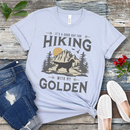 Light blue t-shirt with vintage mountain scene, Golden Retriever silhouette and 'Hiking with my Golden' text, presented on a wooden surface with jeans and shoes.