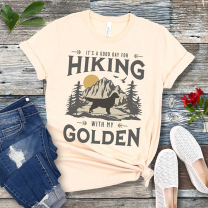 Beige t-shirt with vintage mountain scene, Golden Retriever silhouette and 'Hiking with my Golden' text, presented on a wooden surface with jeans and shoes.