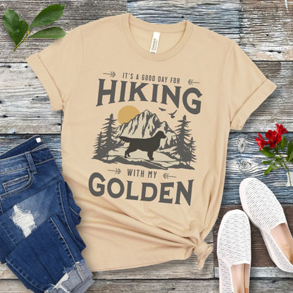 Cream t-shirt with vintage mountain scene, Golden Retriever silhouette and 'Hiking with my Golden' text, presented on a wooden surface with jeans and shoes.