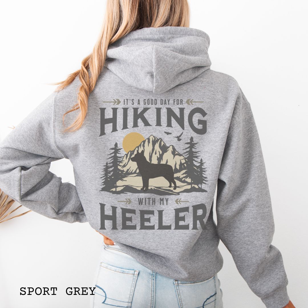 A grey back print hoodie with a graphic print of a Cattle Dog and vintage mountains illustration, and text that reads 'HIKING WITH MY HEELER'. 