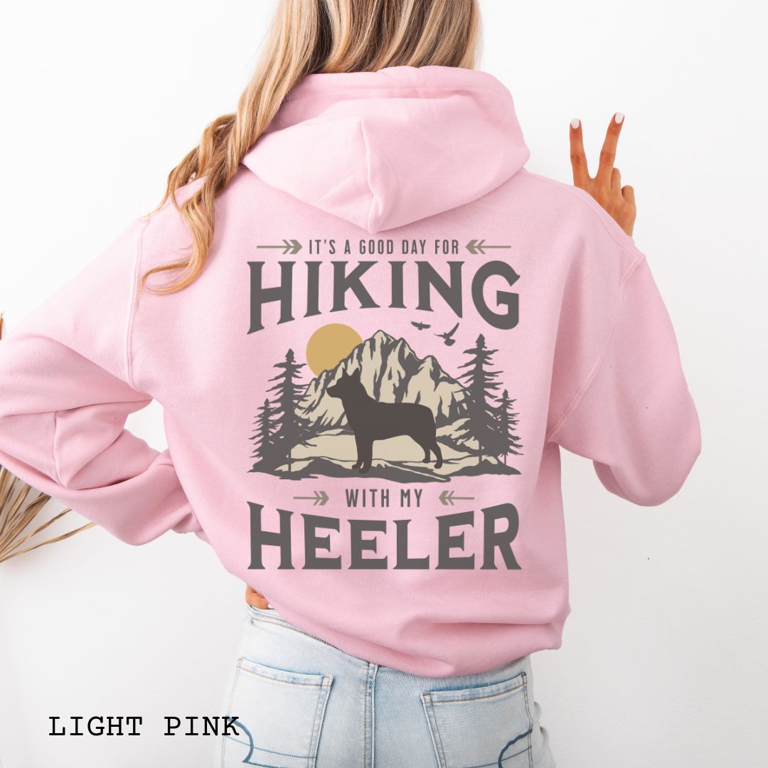 A pink back print hoodie with a graphic print of a Cattle Dog and vintage mountains illustration, and text that reads 'HIKING WITH MY HEELER'. 