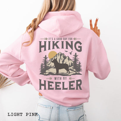 A pink back print hoodie with a graphic print of a Cattle Dog and vintage mountains illustration, and text that reads 'HIKING WITH MY HEELER'. 