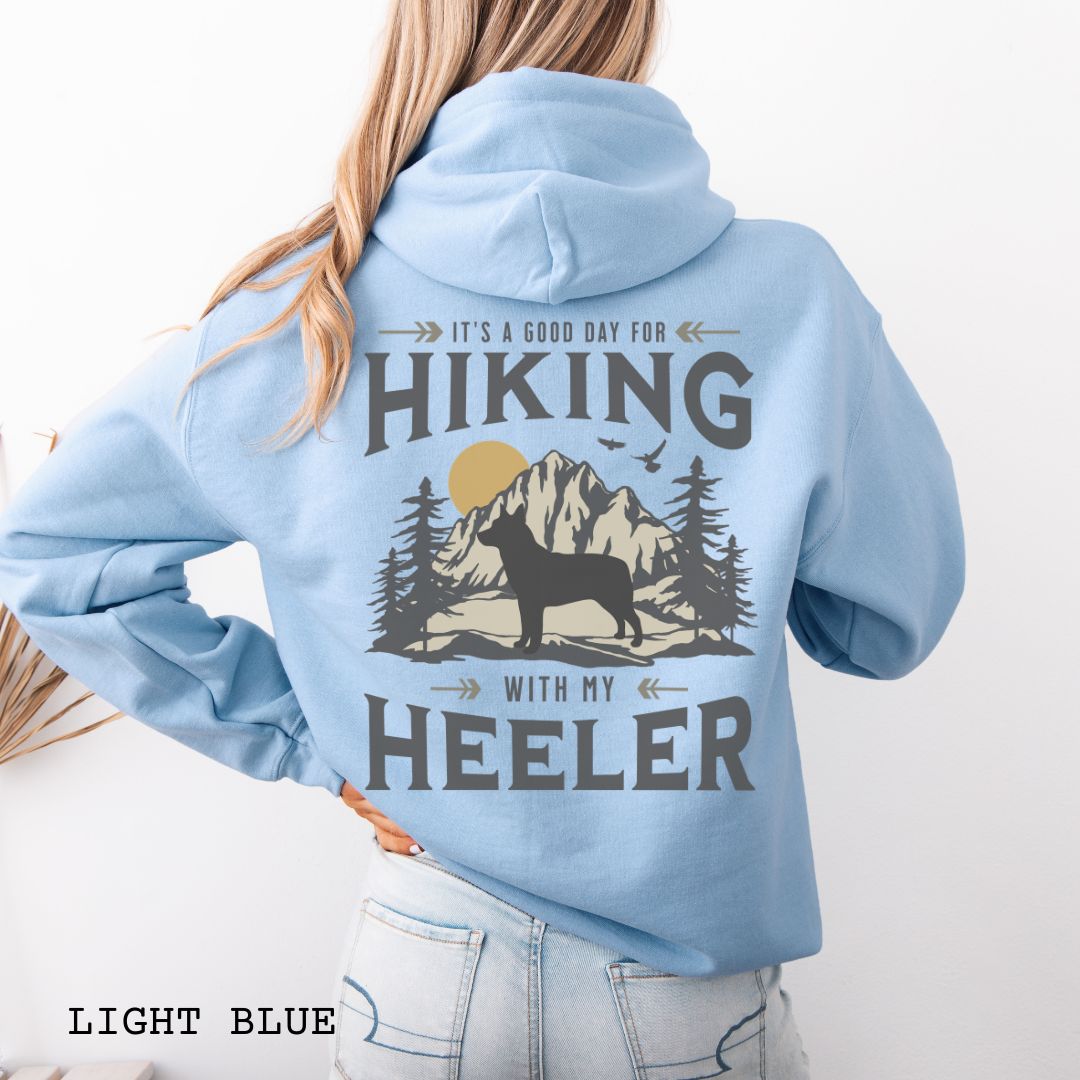 A blue back print hoodie with a graphic print of a Cattle Dog and vintage mountains illustration, and text that reads 'HIKING WITH MY HEELER'. 