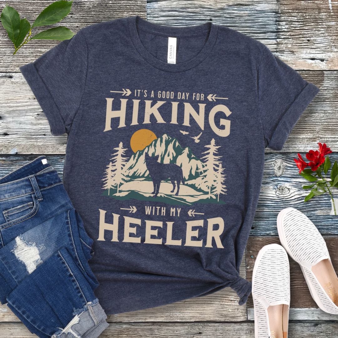 Heather navy t-shirt with a vintage mountain graphic and a silhouette of a Heeler Cattle Dog dog and text 'HIKING with my Heeler, displayed on a wooden surface with denim jeans and white shoes in the background.