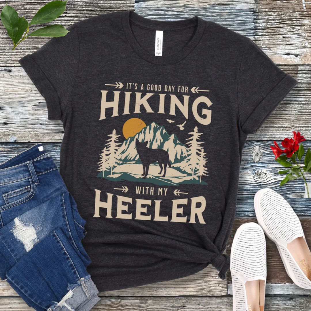 Dark grey t-shirt with a vintage mountain graphic and a silhouette of a Heeler Cattle Dog dog and text 'HIKING with my Heeler, displayed on a wooden surface with denim jeans and white shoes in the background.