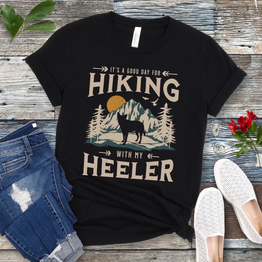 Black t-shirt with a vintage mountain graphic and a silhouette of a Heeler Cattle Dog dog and text 'HIKING with my Heeler, displayed on a wooden surface with denim jeans and white shoes in the background.