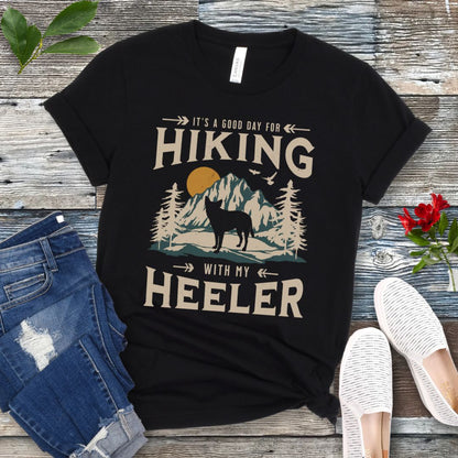 Black t-shirt with a vintage mountain graphic and a silhouette of a Heeler Cattle Dog dog and text 'HIKING with my Heeler, displayed on a wooden surface with denim jeans and white shoes in the background.