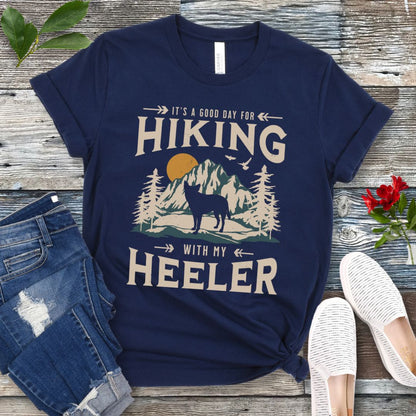 Navy blue t-shirt with a vintage mountain graphic and a silhouette of a Heeler Cattle Dog dog and text 'HIKING with my Heeler, displayed on a wooden surface with denim jeans and white shoes in the background.