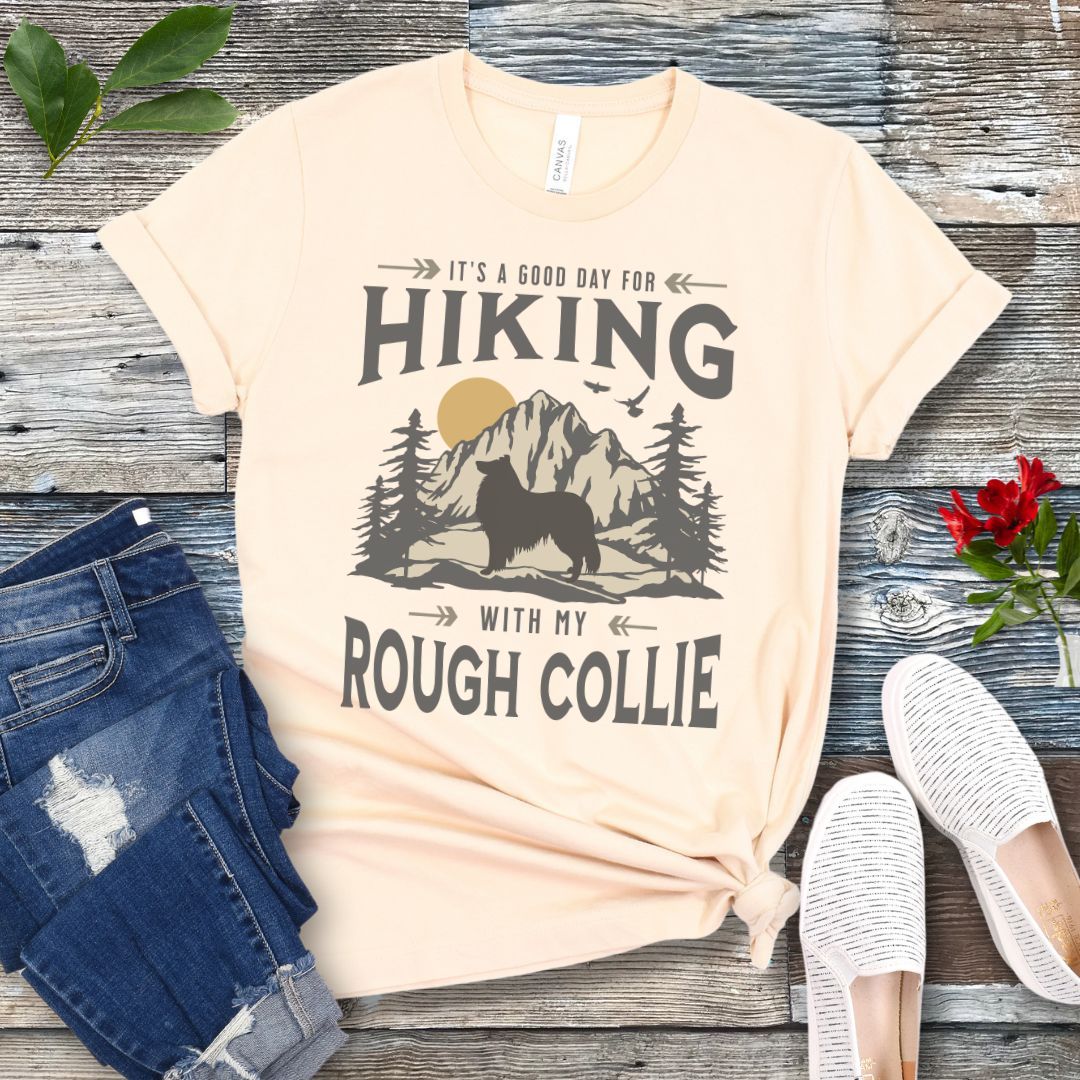 Beige Collie lover t-shirt with vintage mountain scene graphic, a rough collie silhouette and text 'It's a Good Day For Hiking With My Rough Collie', shown on a wooden surface with jeans and shoes.