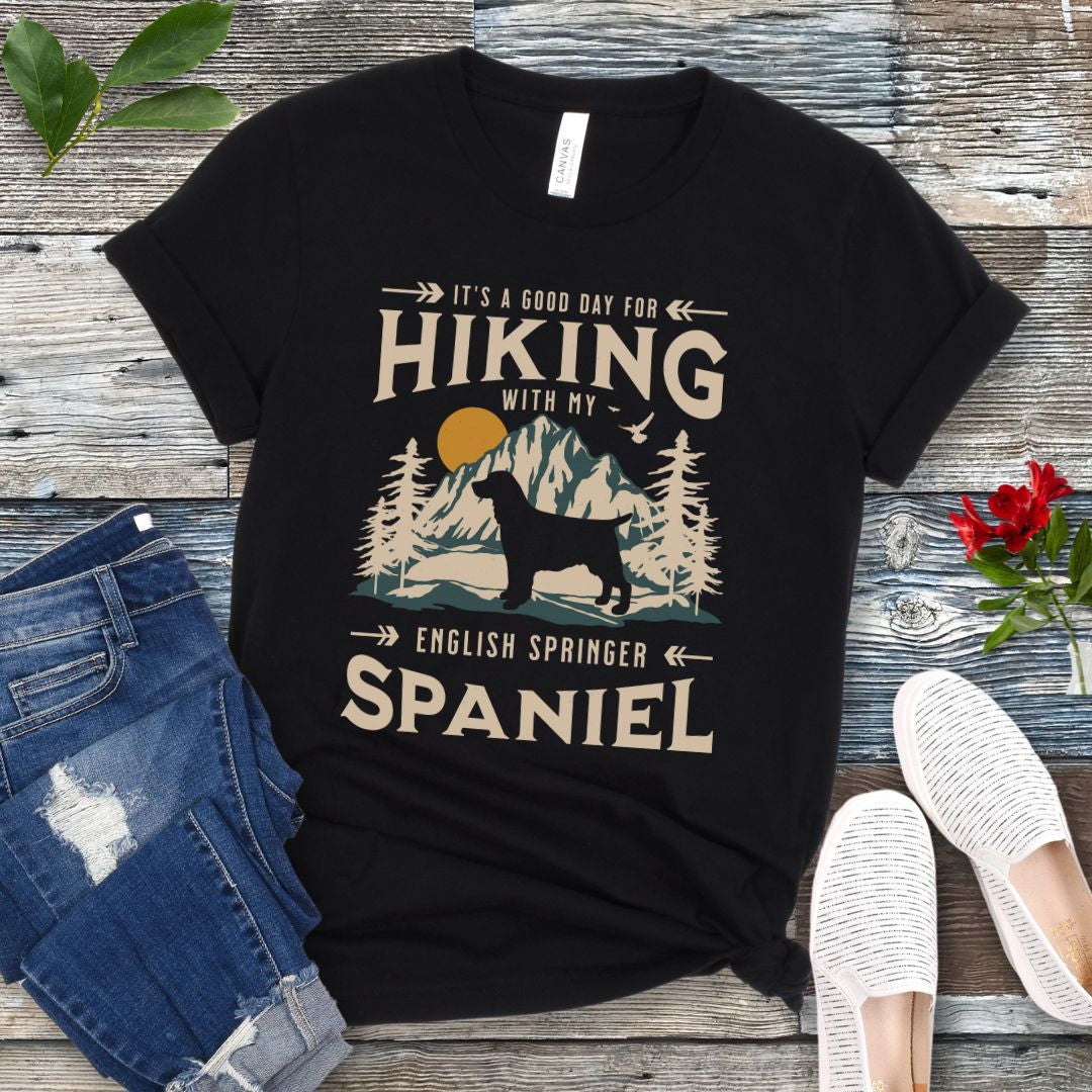 A black t-shirt with a vintage-style graphic print of mountains and a Springer Spaniel and the text 'Hiking with my English Springer Spaniel'. The shirt is presented on a wooden surface with a pair of blue jeans and white shoes in the background.