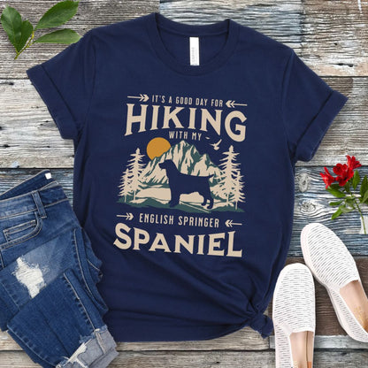 A navy blue t-shirt with a vintage-style graphic print of mountains and a Springer Spaniel and the text 'Hiking with my English Springer Spaniel'. The shirt is presented on a wooden surface with a pair of blue jeans and white shoes in the background.