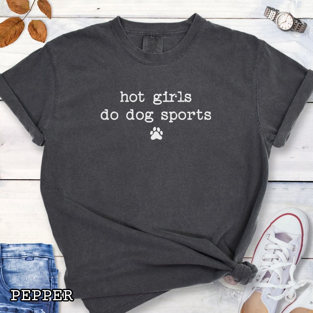 Dark grey Comfort Colors t-shirt for dog agility, nosework, barn hunt, and flyball lovers, with humorous text 'hot girls do dog sports', shown on a wooden surface with jeans and shoes. 