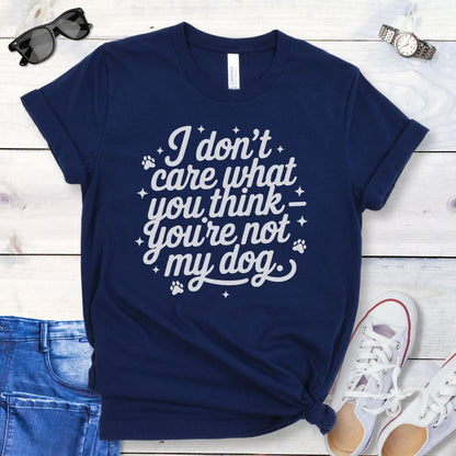 A navy colored dog lover t-shirt with the typography text 'I don't care what you think you're not my dog' printed on it, accented by stars and paw prints, displayed flat with a pair of blue jeans and white shoes.