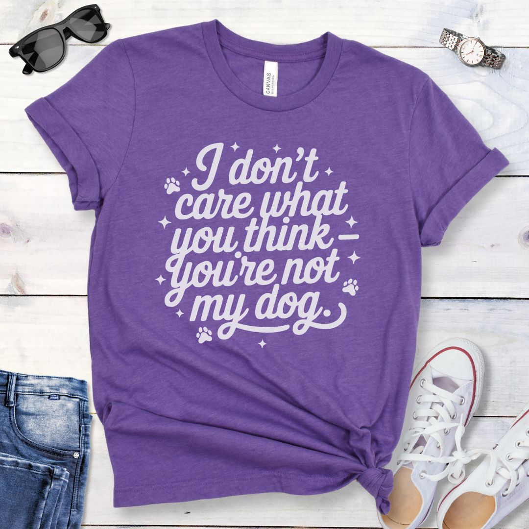 A purple colored dog lover t-shirt with the typography text 'I don't care what you think you're not my dog' printed on it, accented by stars and paw prints, displayed flat with a pair of blue jeans and white shoes.