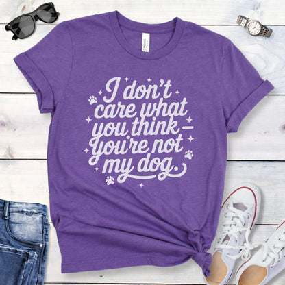 A purple colored dog lover t-shirt with the typography text 'I don't care what you think you're not my dog' printed on it, accented by stars and paw prints, displayed flat with a pair of blue jeans and white shoes.