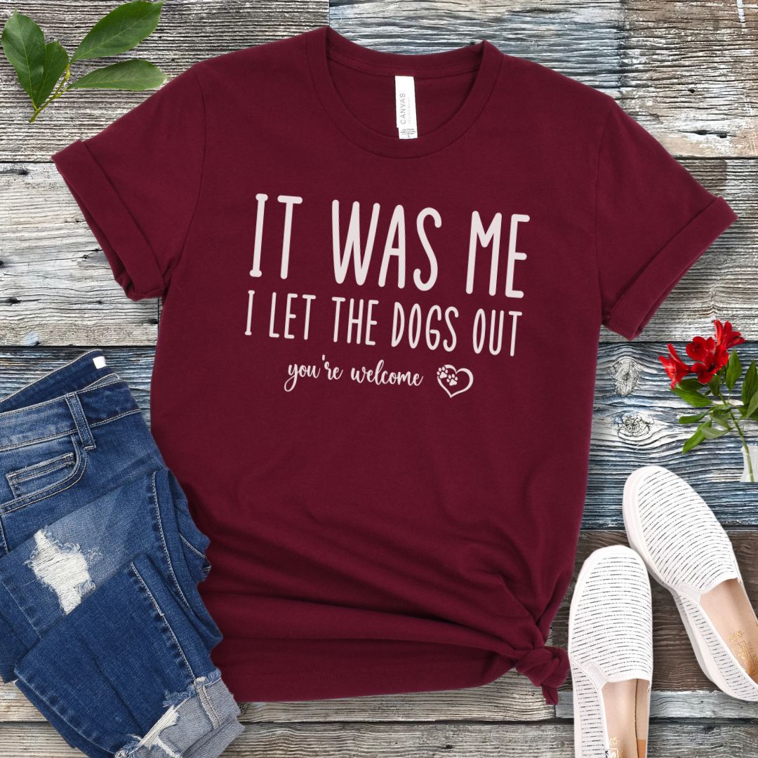 A maroon dog lover t-shirt with the humorous text 'IT WAS ME, I LET THE DOGS OUT, you're welcome' printed in a playful lettering, accented by a heart with paws in it, displayed flat with a pair of blue jeans and white shoes.