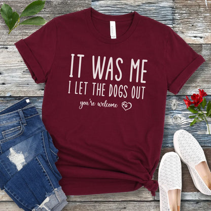 A maroon dog lover t-shirt with the humorous text 'IT WAS ME, I LET THE DOGS OUT, you're welcome' printed in a playful lettering, accented by a heart with paws in it, displayed flat with a pair of blue jeans and white shoes.