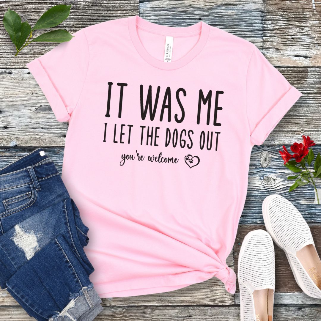A pink dog lover t-shirt with the humorous text 'IT WAS ME, I LET THE DOGS OUT, you're welcome' printed in a playful lettering, accented by a heart with paws in it, displayed flat with a pair of blue jeans and white shoes.