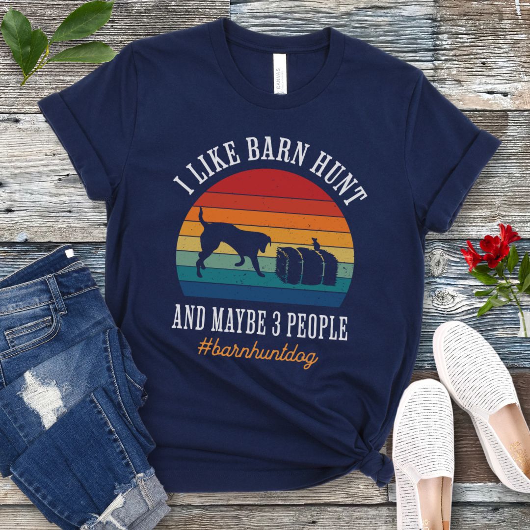Navy dog sports t-shirt with colorful graphic and the text 'I Like Barn Hunt And Maybe 3 People' on a wooden surface with jeans and shoes.