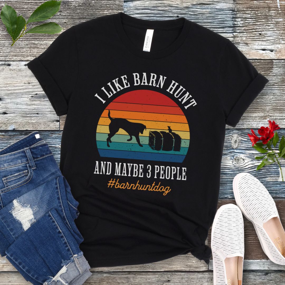 Black dog sports t-shirt with colorful graphic and the text 'I Like Barn Hunt And Maybe 3 People' on a wooden surface with jeans and shoes.