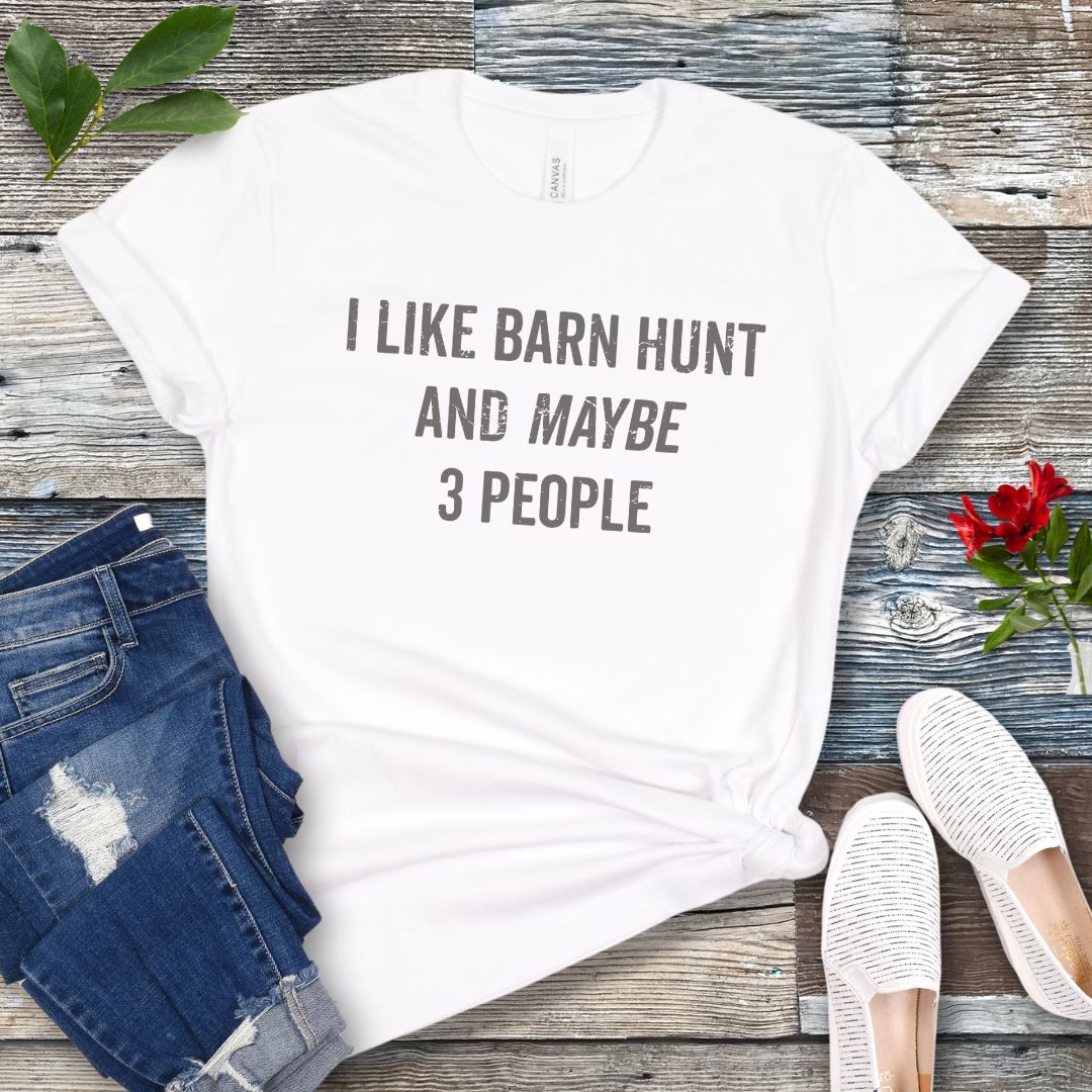 White t-shirt with text 'I LIKE BARN HUNT AND MAYBE 3 PEOPLE' on a wooden surface with jeans and shoes.