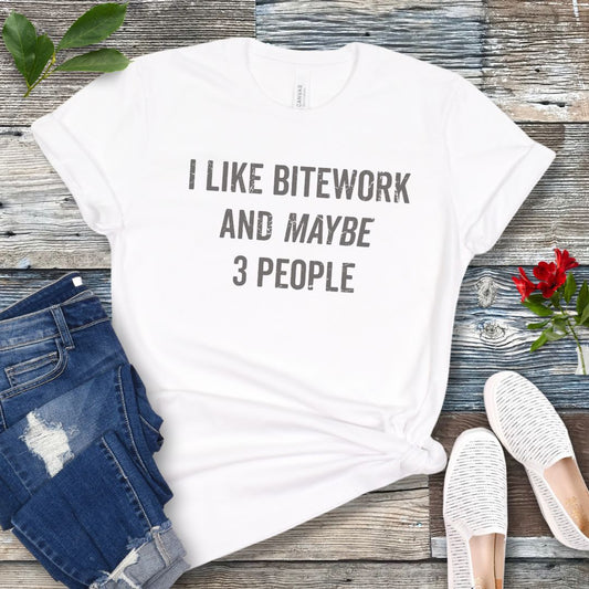 White IGP/IPO t-shirt with 'I LIKE BITEWORK AND MAYBE 3 PEOPLE' text on a wooden surface with jeans and shoes.